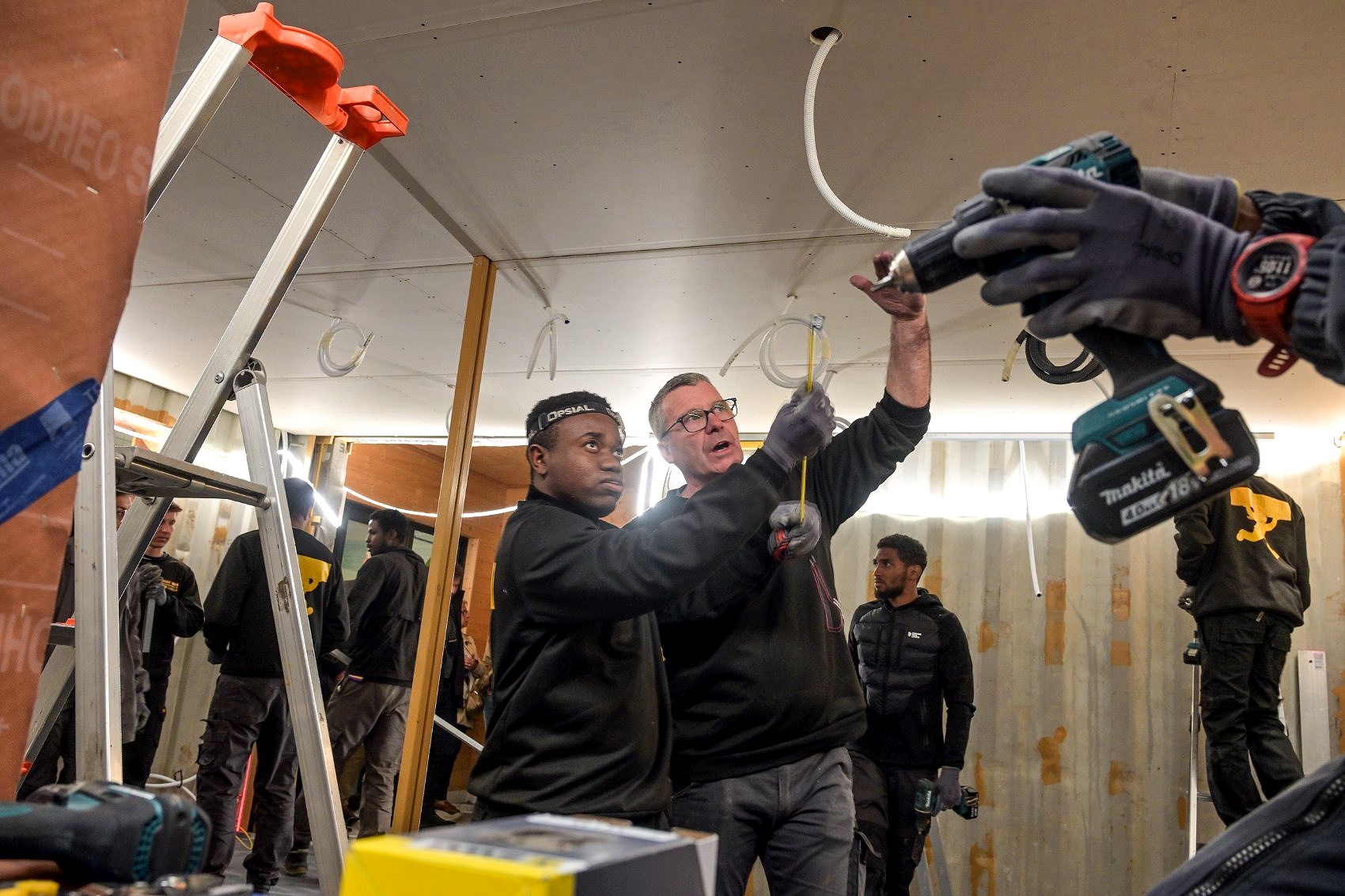 Un formateur de Forma Cargo guide un apprenant lors de l'installation des réseaux au plafond d'un module, entourés d'autres apprenants, dans les ateliers 123modules